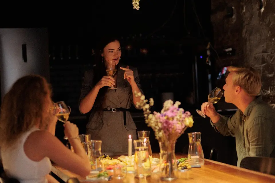 Person standing and speaking with a glass in hand to two others sipping wine, with flowers on the table