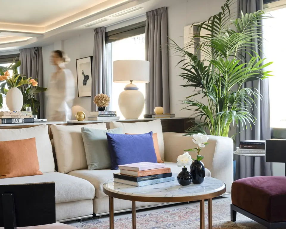 Cream sofa with colourful cushions and a round marble coffee table beside a palm and windows with grey curtains