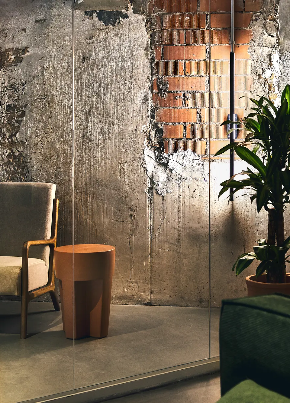 Raw concrete wall with exposed brick behind glass, next to a wooden armchair, a terracotta stool and a potted plant