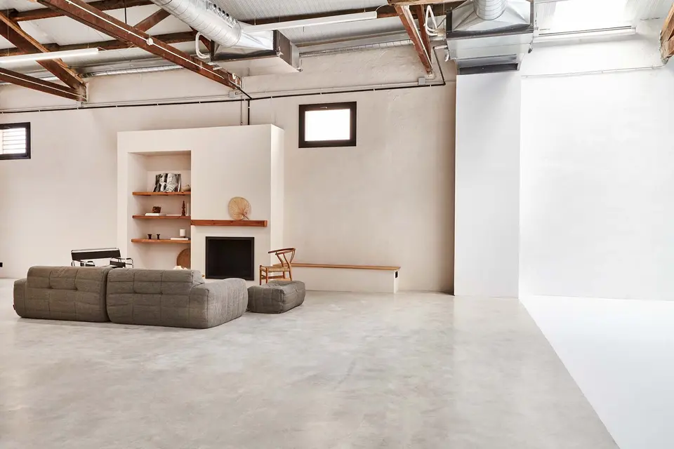 Open room with a polished concrete floor, a grey sofa facing a fireplace with wooden shelves and a white studio backdrop