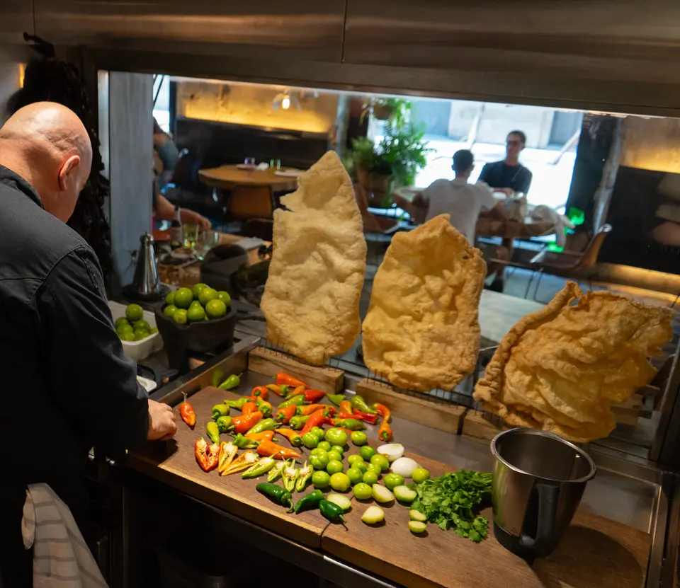 Cook seen from behind at a board of chilies and limes, with three large fried crisps above and diners in the background