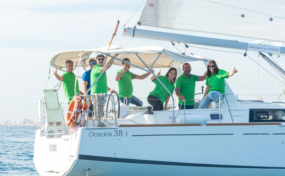 Crew in green polo shirts waving and giving thumbs up from a sailboat stern, next to an orange lifebuoy