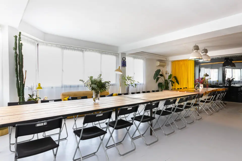 Long plank wood table with black folding chairs on both sides, with a tall cactus and sheer-curtained windows