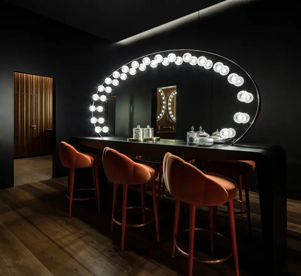 Large oval mirror ringed with bulbs above a black counter with three terracotta upholstered stools, in a dark-walled room