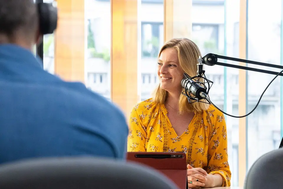 Smiling person at a studio microphone on a boom arm, with another person blurred in the foreground