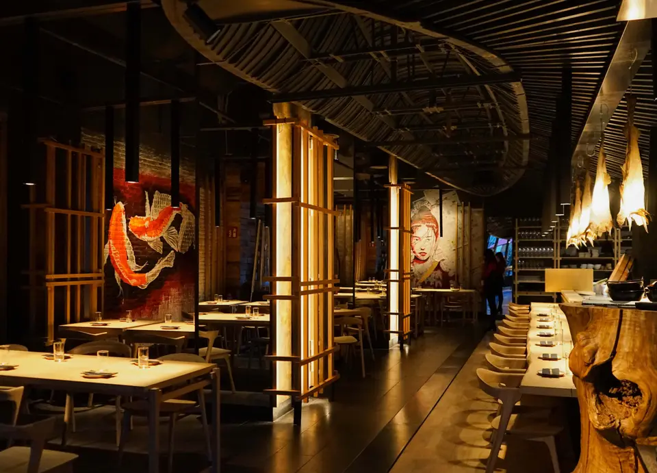 Long dining room with wooden tables, lit slatted pillars and a fish mural at the back, in warm dim light