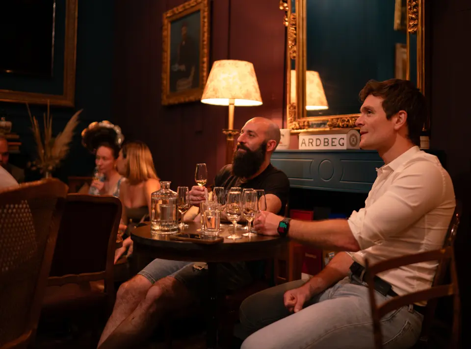 Group seated around a table with glasses in a lounge with a gilded mirror and a lit lamp