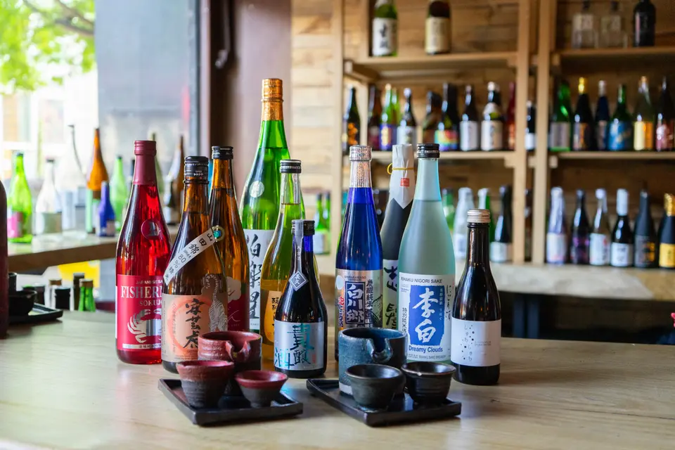 Colorful sake bottles on a wooden counter, with small ceramic cups on black trays and shelves behind