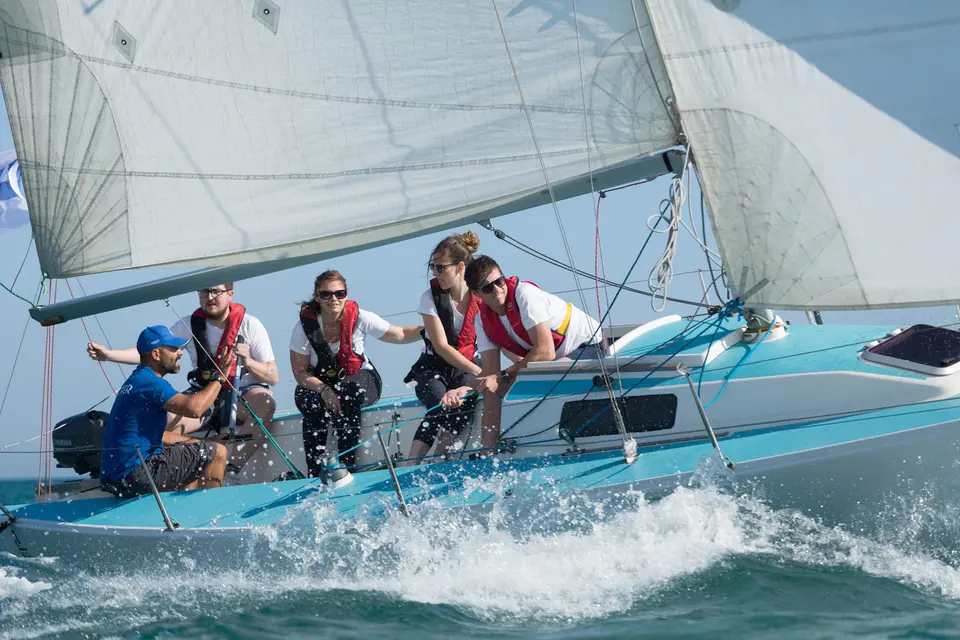 Crew in life jackets on the rail of a light blue sailboat throwing up spray as it sails