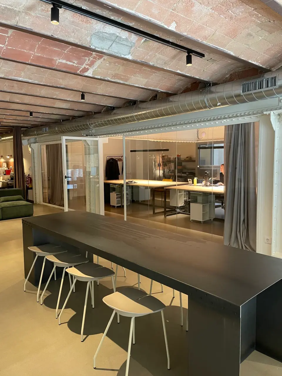 Tall black table with four stools under an exposed brick ceiling, with glass-walled offices at the back of the room