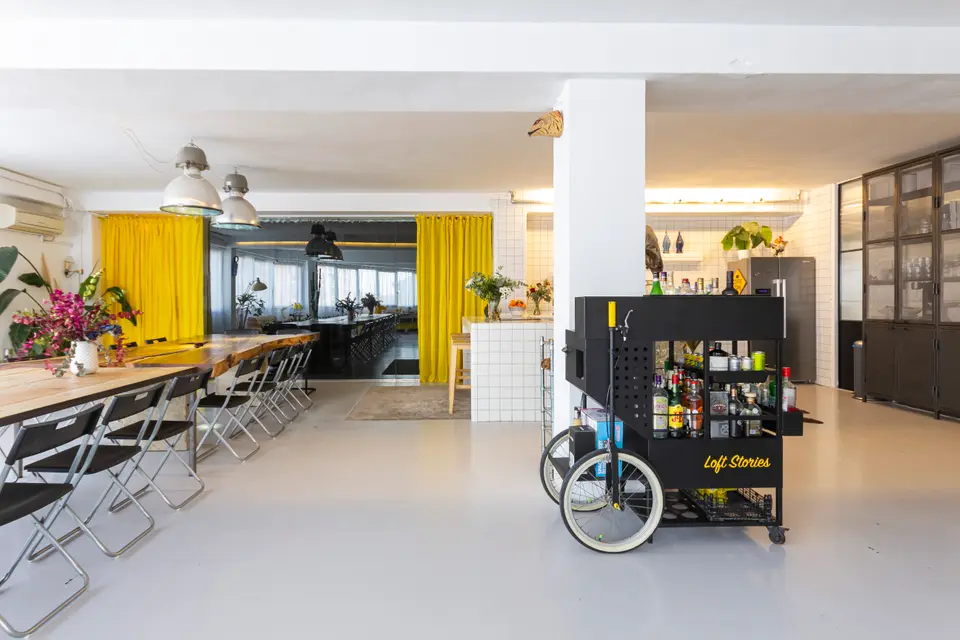 Black bar cart with bottles and the Loft Stories lettering, in front of a long table and yellow curtains at the back