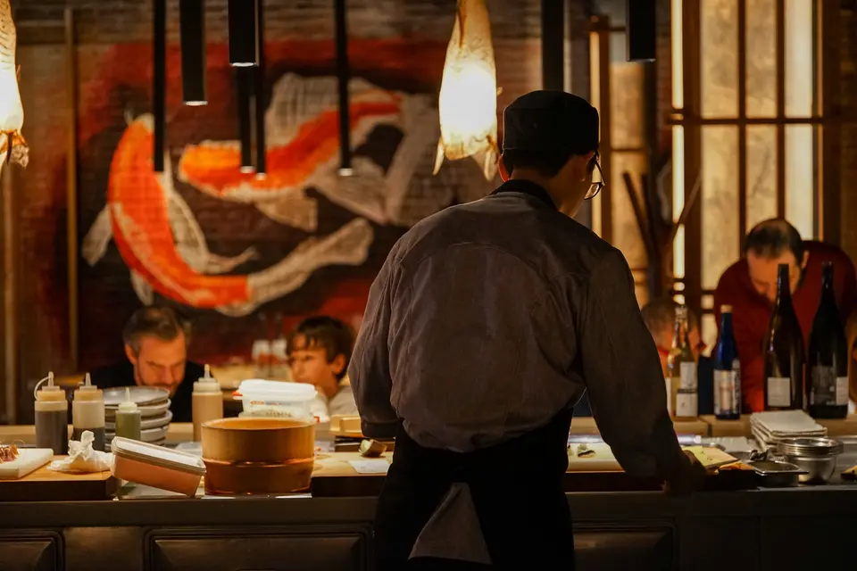 Cook seen from behind at an open kitchen counter, with two more behind and a mural of orange koi fish
