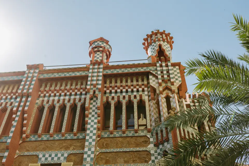 Tiled and brick façade with two turrets seen from below, with palm leaves on the right