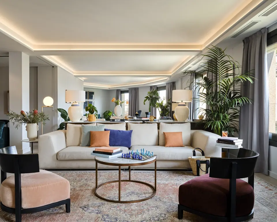 Living area with a long cream sofa, two armchairs and a brass coffee table with a chess set, under cove lighting